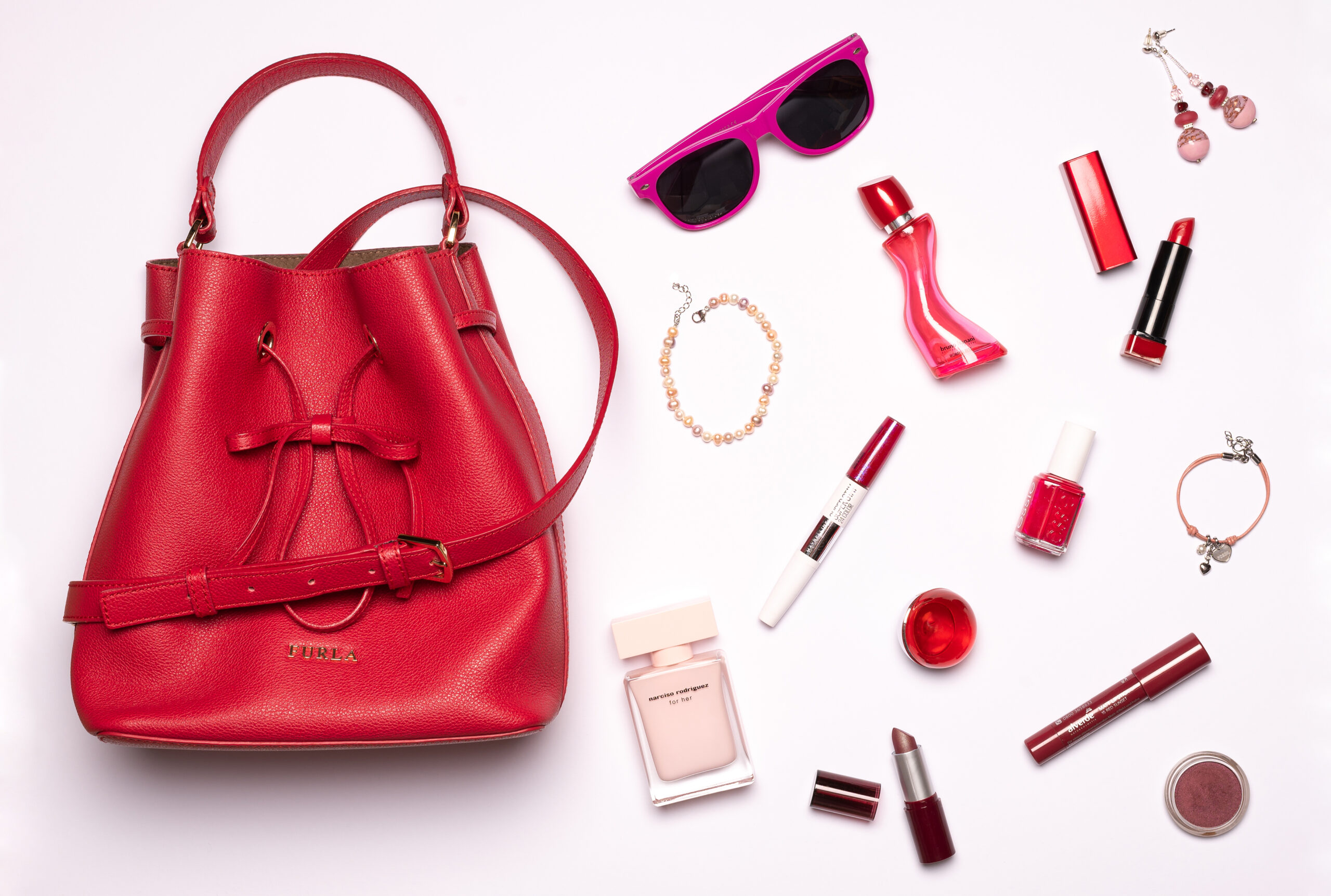 Produktfotografie: Pinke Handtasche links mit Sonnenbrille, Schmuck, Make-up und Parfum rechts auf weißem Hintergrund